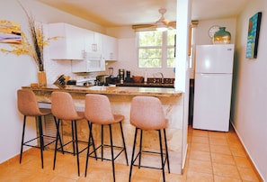 Una cocina con armarios blancos, encimera de granito, cuatro taburetes de bar, un refrigerador y una ventana con vistas a árboles.