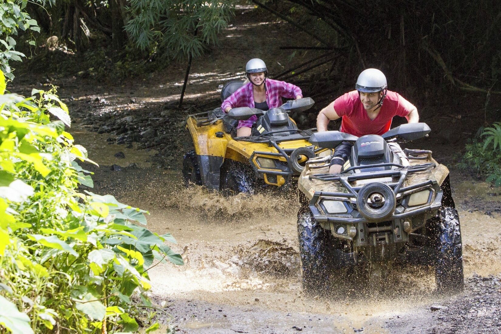 Zwei Personen fahren mit Quads auf einem schlammigen Pfad durch ein bewaldetes Gebiet.