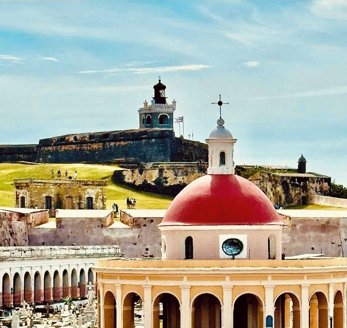 A historical fort with a red dome and a clock tower, surrounded by a fortified wall and a courtyard with arches.