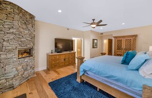 A bedroom with a stone fireplace, a bed with blue bedding, a wooden dresser, and a ceiling fan.