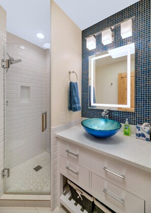 A bathroom with a glass shower enclosure, a white vanity with a blue glass sink, and a mirror with blue mosaic tiles.