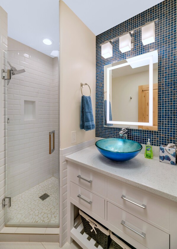 A bathroom with a glass shower enclosure, a white vanity with a blue glass sink, and a mirror with blue mosaic tiles.