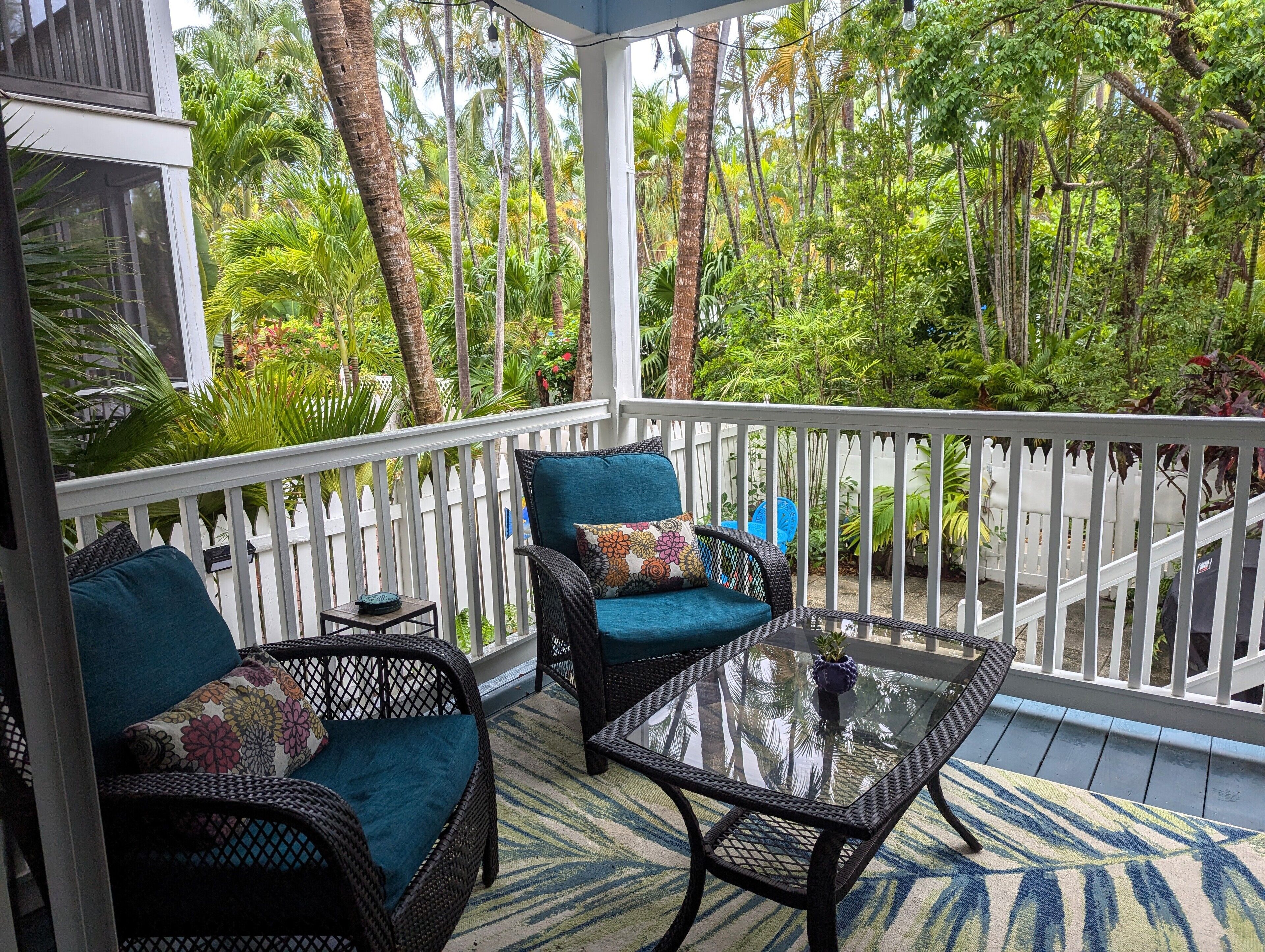 A covered outdoor area with wicker furniture and a glass table, surrounded by a white fence and lush greenery.