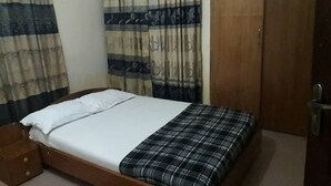 Un lit simple avec un drap blanc et une couverture à carreaux, une table de chevet en bois et une porte avec une poignée.