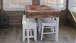 Een houten blokhut van binnen, met een eettafel voor vier personen, witte stoelen en grote ramen met zonwering.