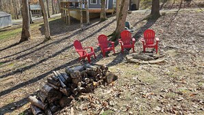 Een chalet met terras, omringd door bomen en een vuurplaats met rode stoelen.