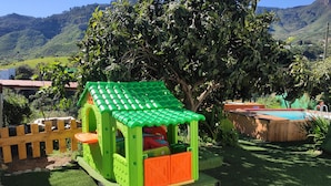 A green and orange playhouse with a red slide, surrounded by a wooden fence, in a backyard with a pool and a mountain backdrop. - Cozy house in a rural setting (tenteniguada)