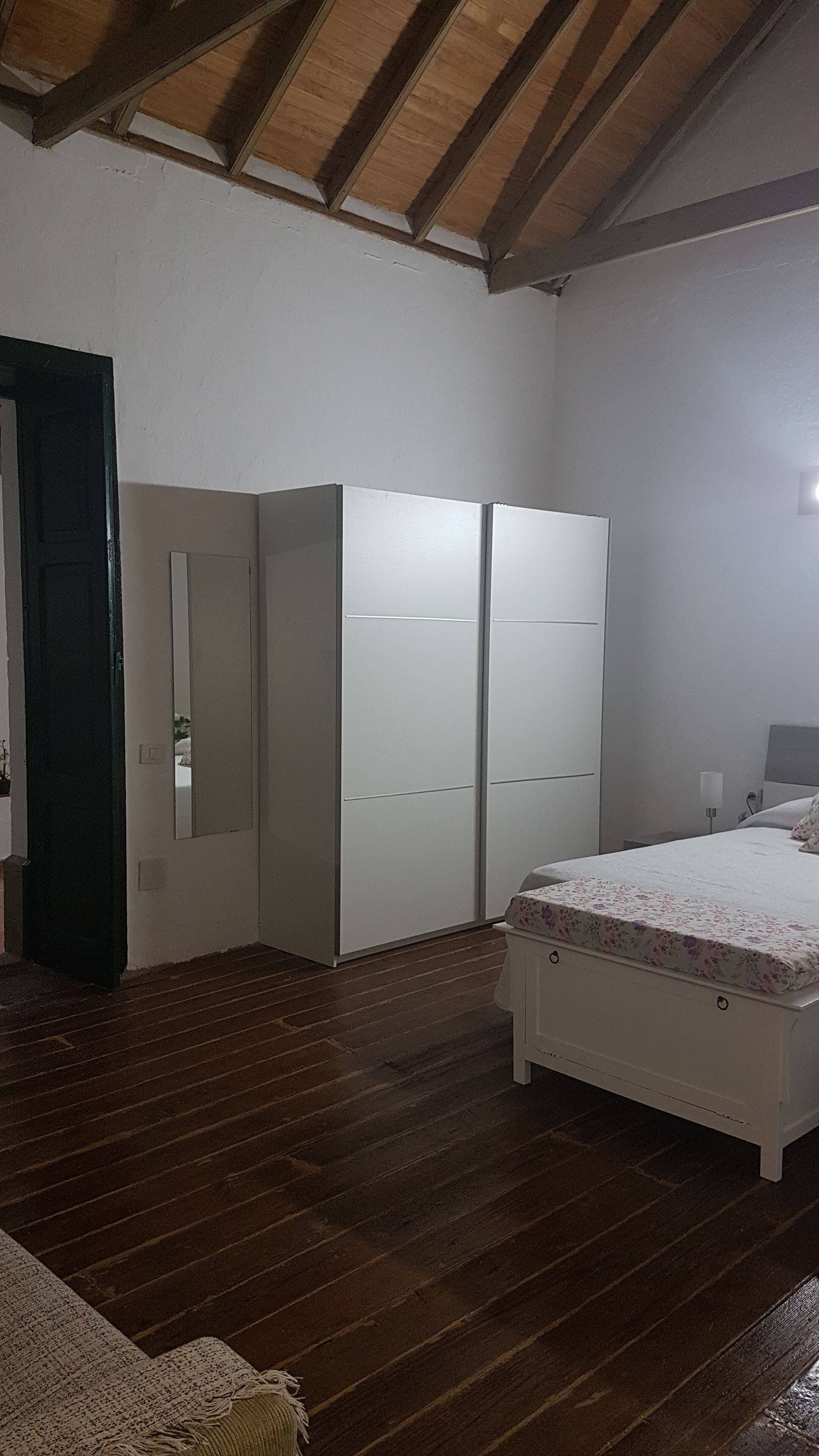 A bedroom with a wooden floor, a bed with a floral bedspread, a wardrobe, and a mirror.