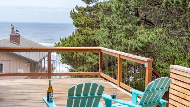 A wooden deck with two turquoise Adirondack chairs, a bottle of wine, and a glass. The deck overlooks a beach and ocean, with a building in the background. - hot tub, ocean views and wood burning fireplace (Tillamook)