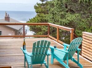 A wooden deck with two turquoise Adirondack chairs, a bottle of wine, and a glass. The deck overlooks a beach and ocean, with a building in the background.