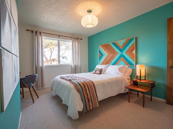 A bedroom with a bed, a nightstand, a lamp, a chair, a window with curtains, and a wall with a geometric painting.