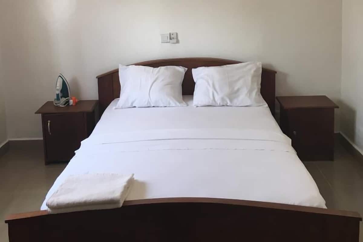 Habitación de hotel sencilla y limpia con una cama individual, dos almohadas blancas, un cabecero de madera, una mesita de noche con un planchador y una pequeña mesa.