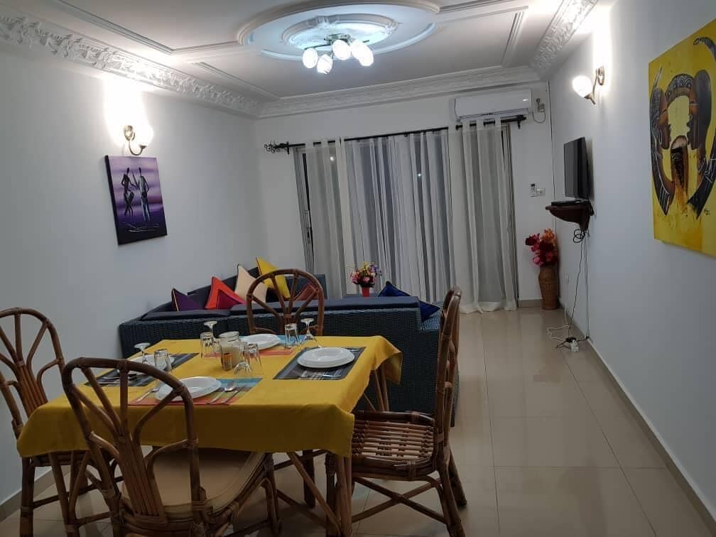 A dining area with a table set for four, surrounded by wicker chairs, a sofa with cushions, and a large window with curtains.