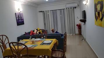 A dining area with a table set for four, surrounded by wicker chairs, a sofa with cushions, and a large window with curtains.