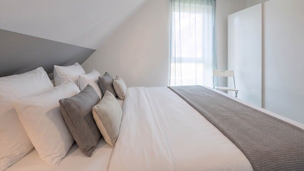Een slaapkamer met een bed, kussens, een raam met gordijnen, een stoel en een kledingkast.