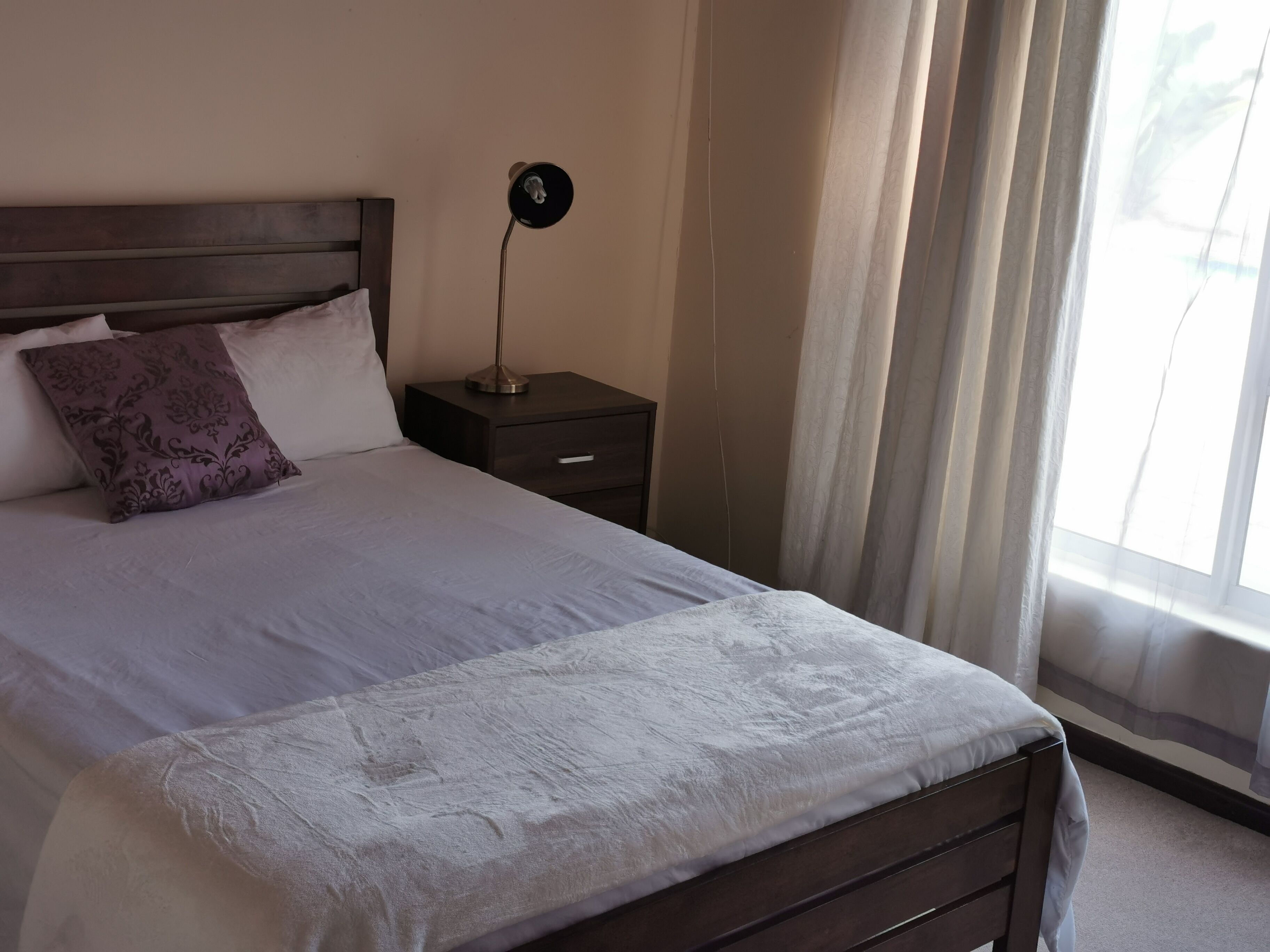 A neatly made bed with a white blanket and a decorative pillow, a bedside table with a lamp, and a window with curtains.