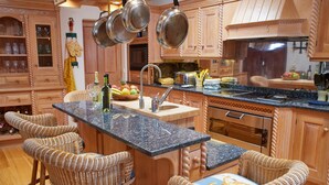 Una cocina con armarios de madera, encimera de granito, pileta de acero inoxidable y horno empotrado. Hay sillas de mimbre alrededor de la isla y una botella de vino en la encimera.