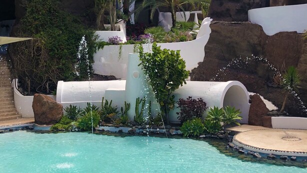 Una piscina con un telón de fondo de roca natural, rodeada de diversas plantas y una estructura blanca con fuente de agua.