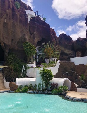 A pool with a natural rock backdrop, surrounded by various plants and a white structure with a water feature. - Luxurious Oceanfront Villa with Panorama View and Private Pool (Costa Teguise)