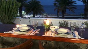Mesa para dos con platos, vasos y una vela, con vista a una playa con palmeras y el océano.