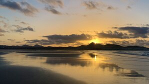 Una playa con un atardecer, montañas al fondo y agua tranquila que refleja el cielo.