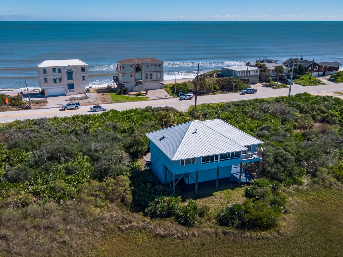 A coastal house with a blue roof, surrounded by dense vegetation and a view of the ocean.