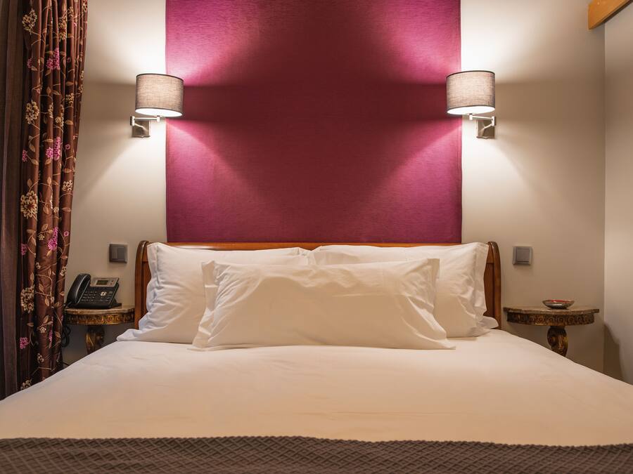 Une chambre d’hôtel avec un grand lit, deux lampes de chevet, un téléphone, une petite table avec un bol, et un rideau décoratif.