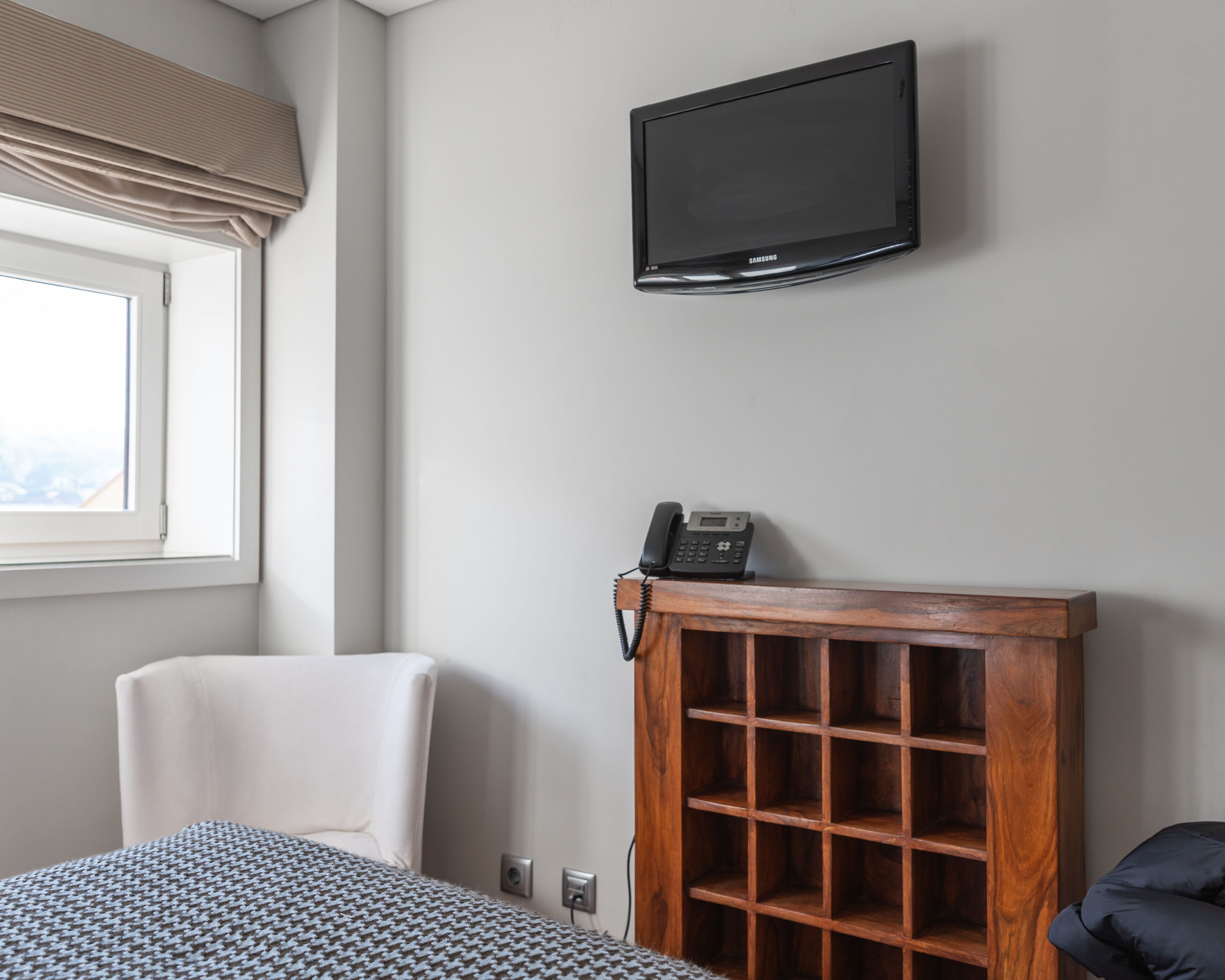 A hotel room with a bed, a chair, a television mounted on the wall, and a wooden cabinet with a telephone.