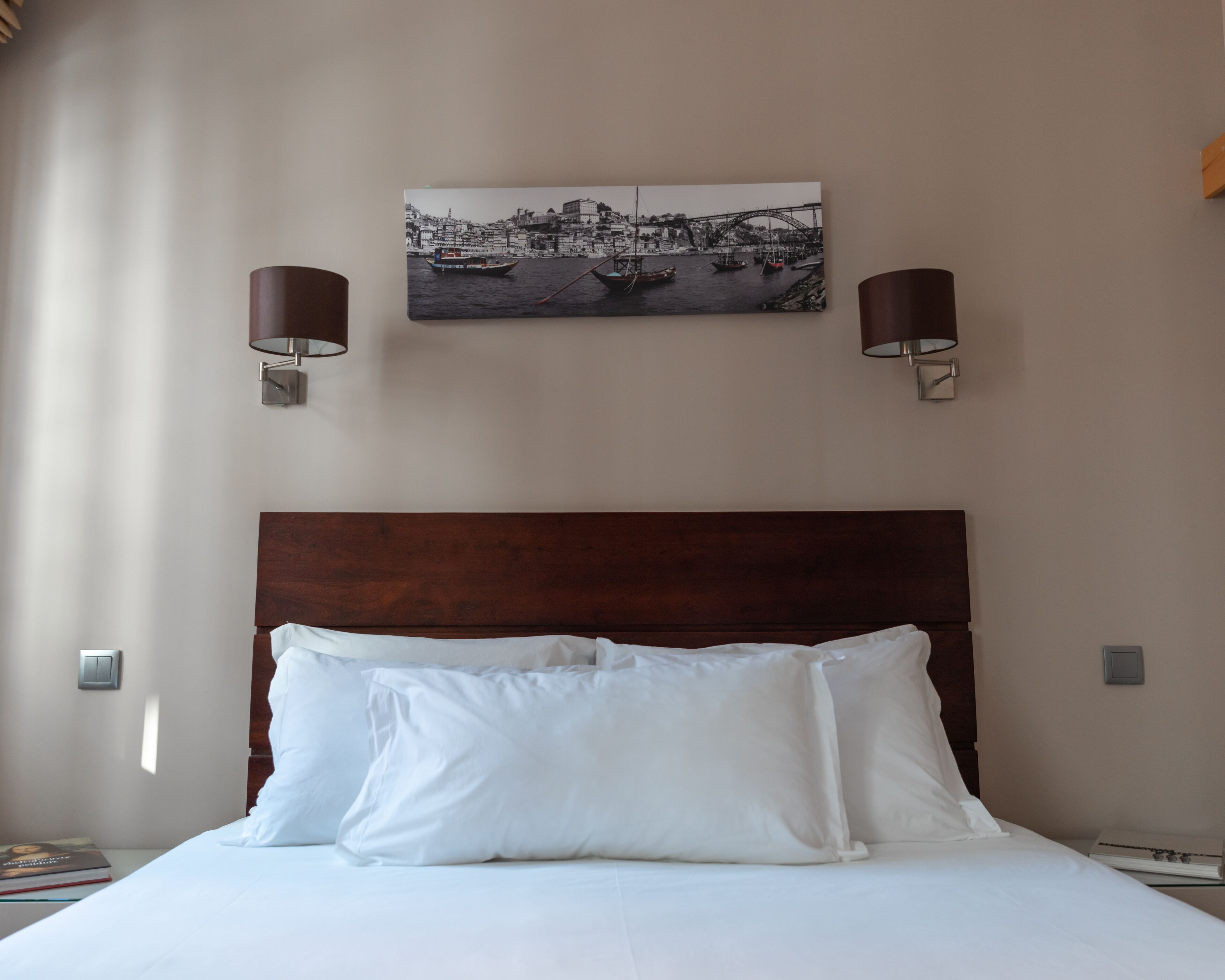A hotel room with a wooden headboard, white bedding, and a cityscape painting above the bed.
