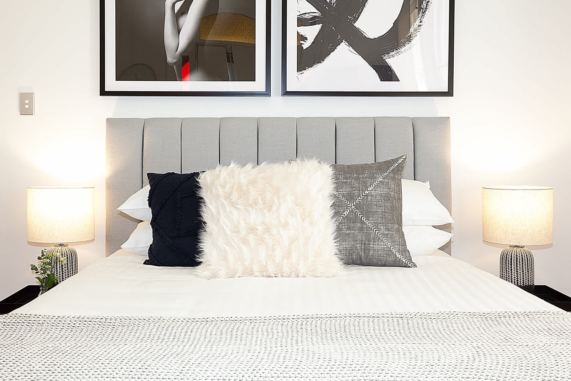 A neatly made bed with a white duvet, flanked by two bedside lamps and three decorative pillows. Above the bed, there are two framed artworks on the wall.