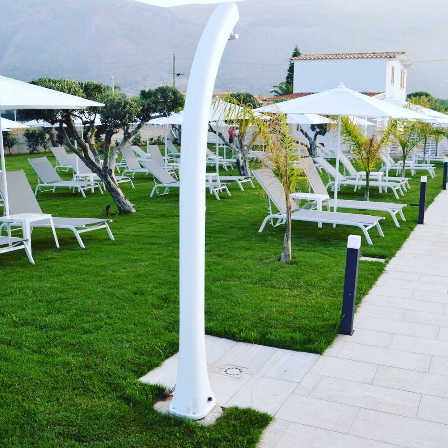 A well-maintained outdoor lounge area with white sun loungers and umbrellas, a paved walkway, and a backdrop of greenery and a building.