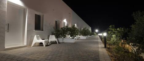 A paved walkway leading to a building with white walls and doors, benches, and potted plants along the path.