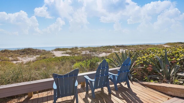 Una terraza de madera con sillas azules da a una playa con dunas y vegetación.