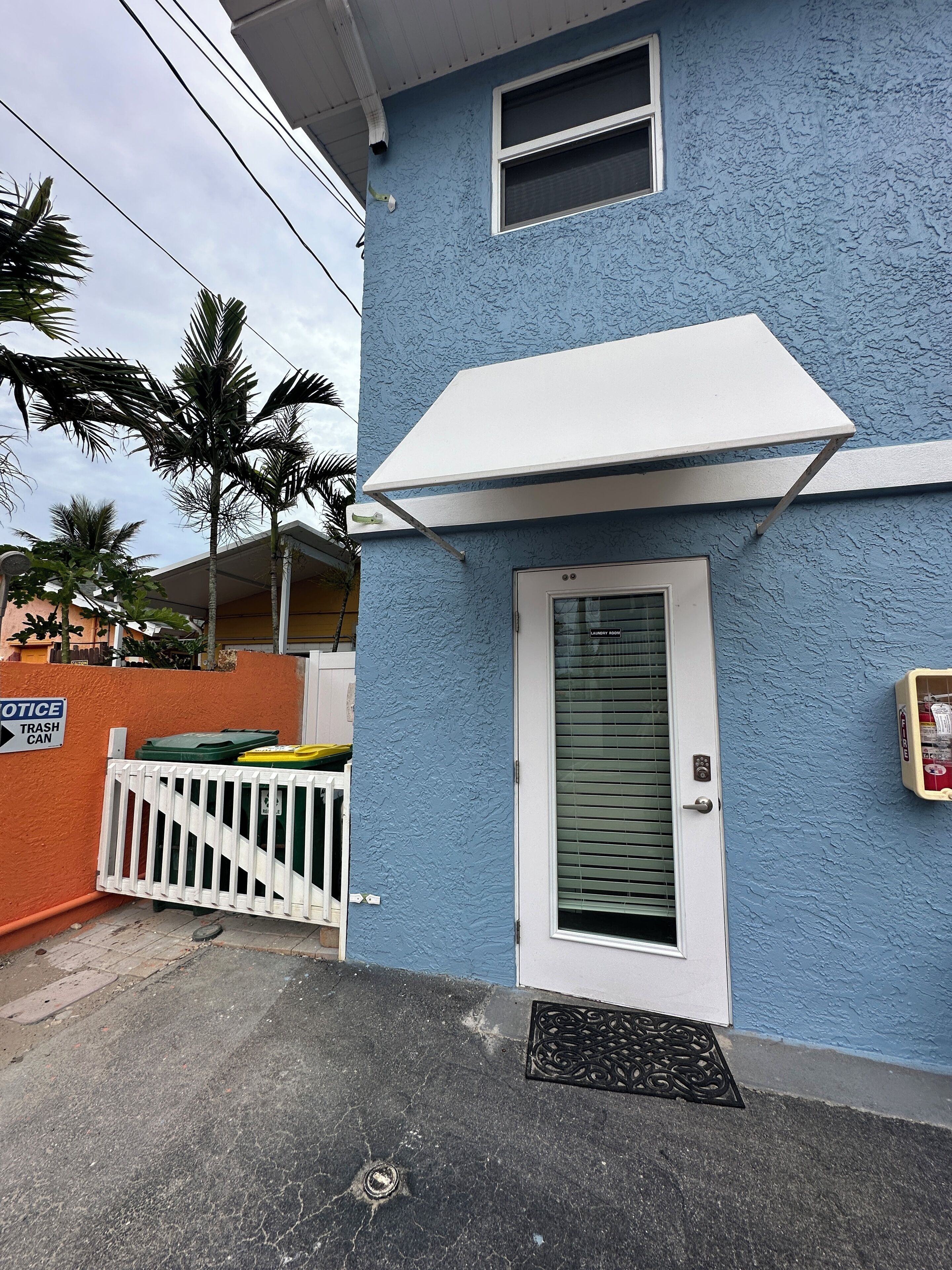 A blue building with a white door and window, a white awning, and a trash can outside.