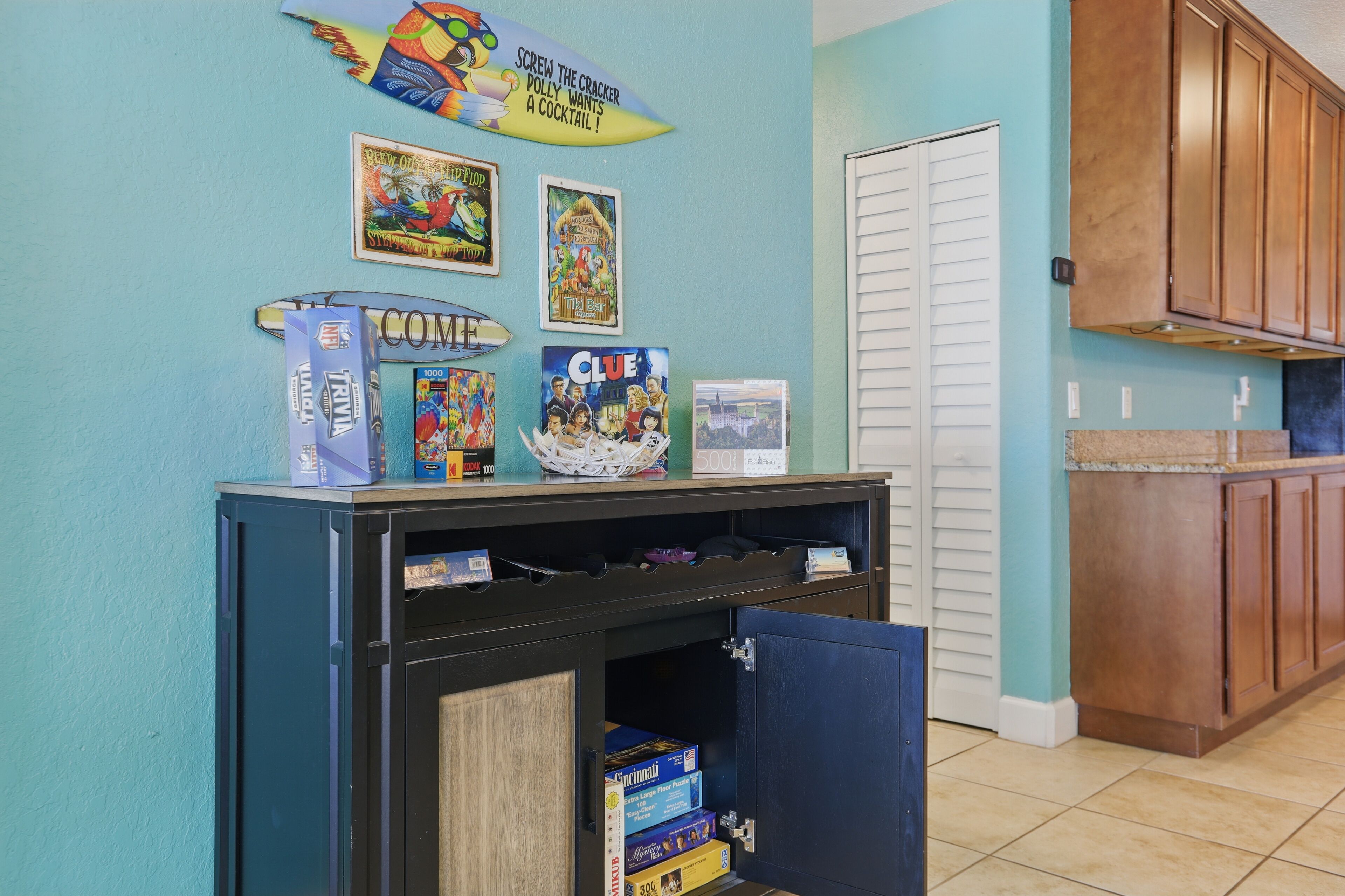 A kitchen with teal walls, wooden cabinets, and a countertop. There are several board games on a shelf, including 'Trivial Pursuit' and 'Clue'.