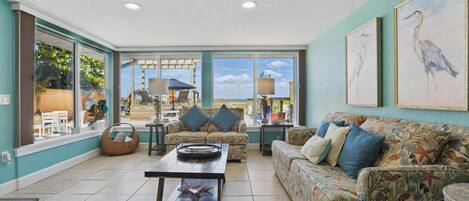 A living room with a sofa, coffee table, and chairs. There are large windows and a view of the outside.