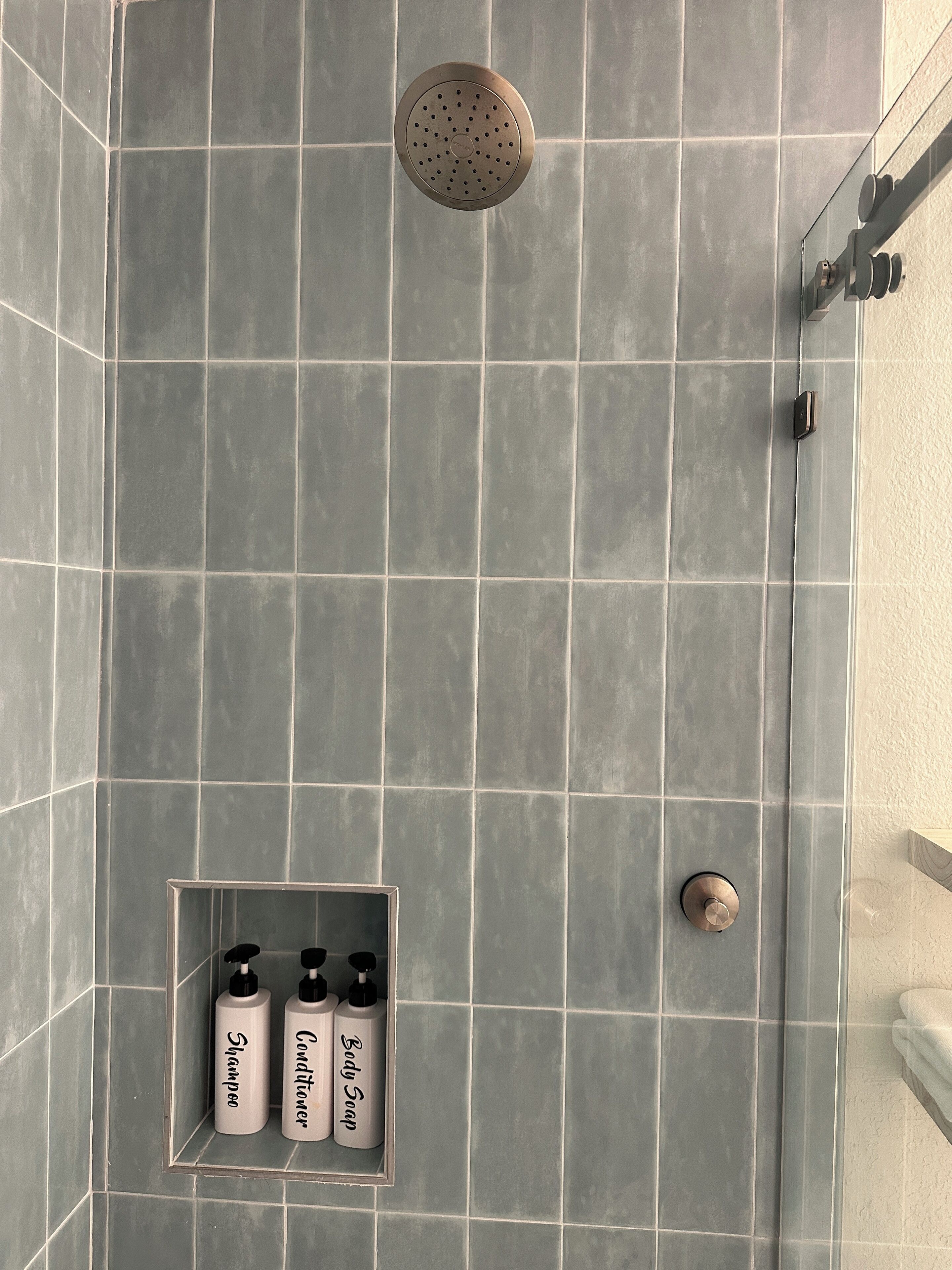 Une salle de bain moderne avec des carreaux gris, une pomme de douche et trois flacons de shampoing, d’après-shampoing et de savon pour le corps.