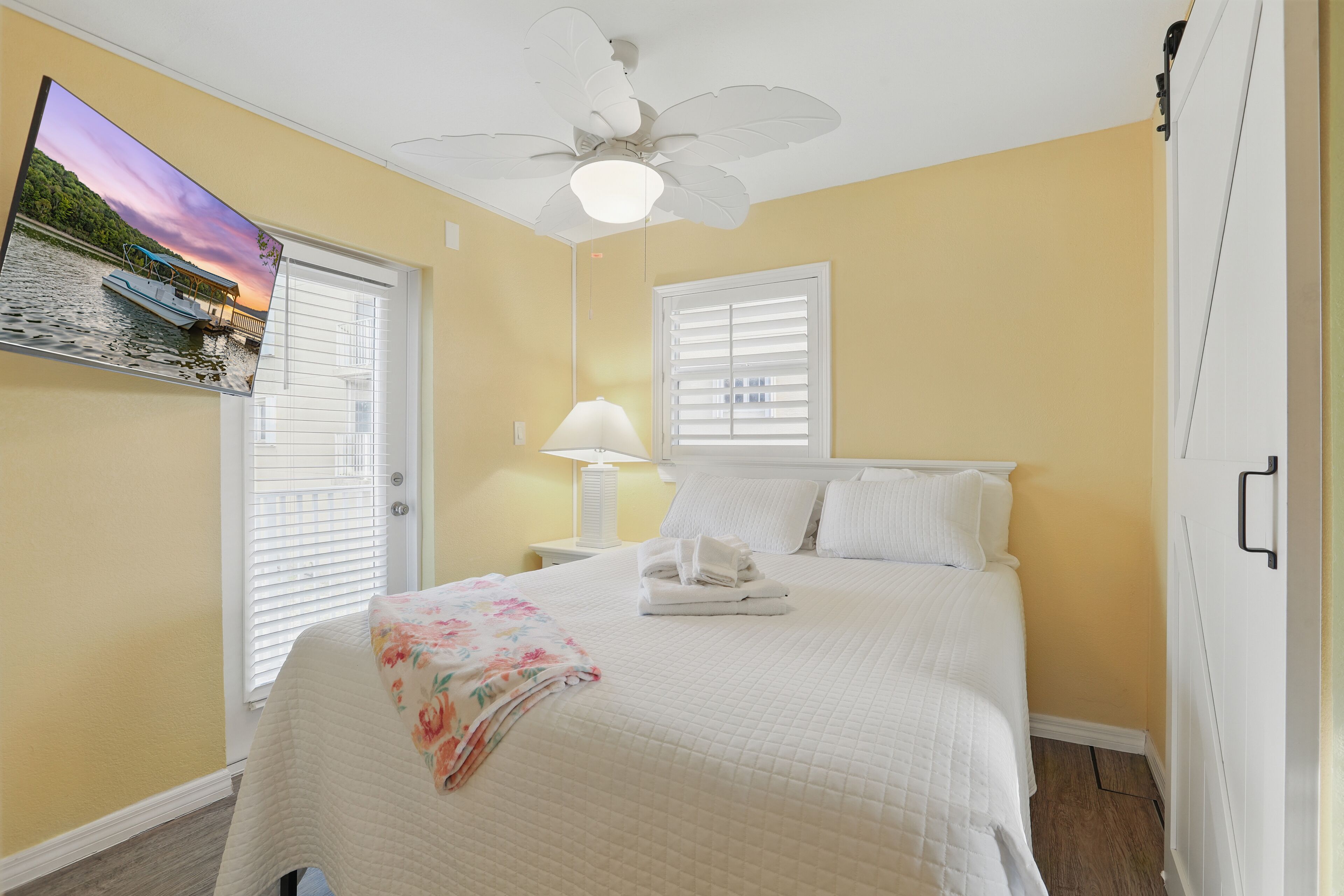 Une chambre à coucher avec un lit, un ventilateur de plafond, un téléviseur fixé au mur et une fenêtre avec des stores.