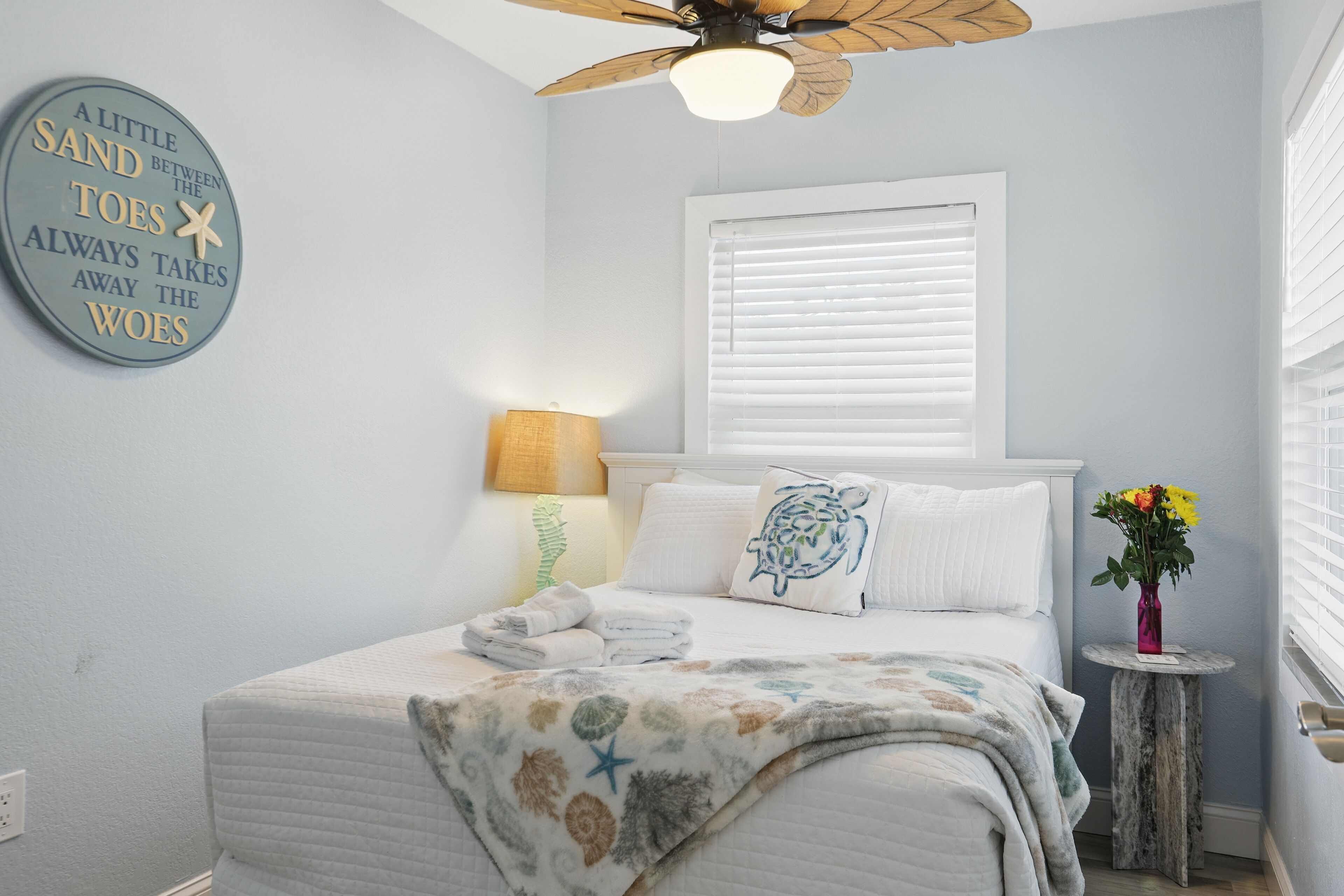A bedroom with a bed, a ceiling fan, a window with blinds, a bedside lamp, a decorative wall clock, and a vase with flowers.