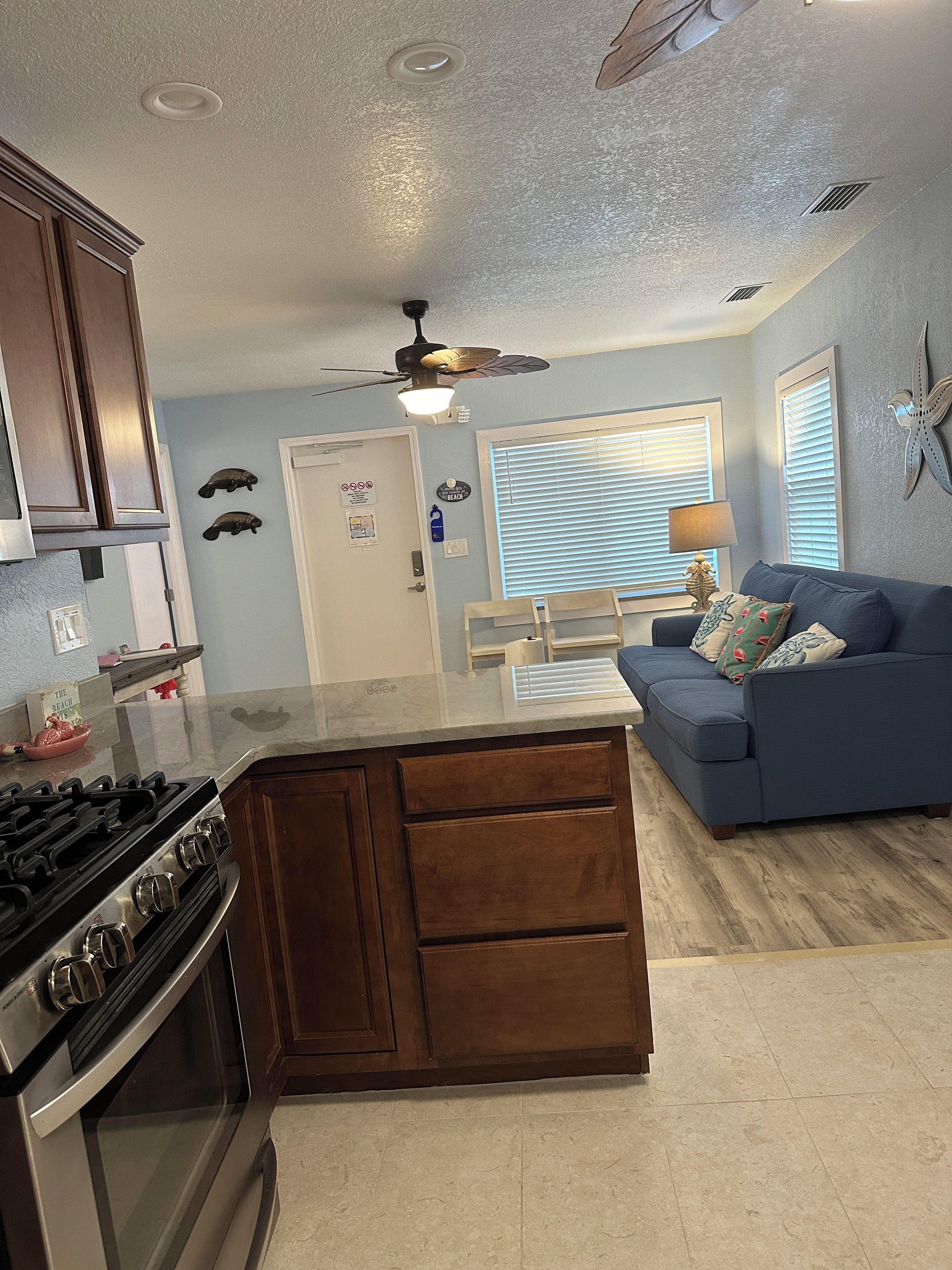 A kitchen with a stove, oven, and wooden cabinets. There is a blue couch and a ceiling fan in the living area.