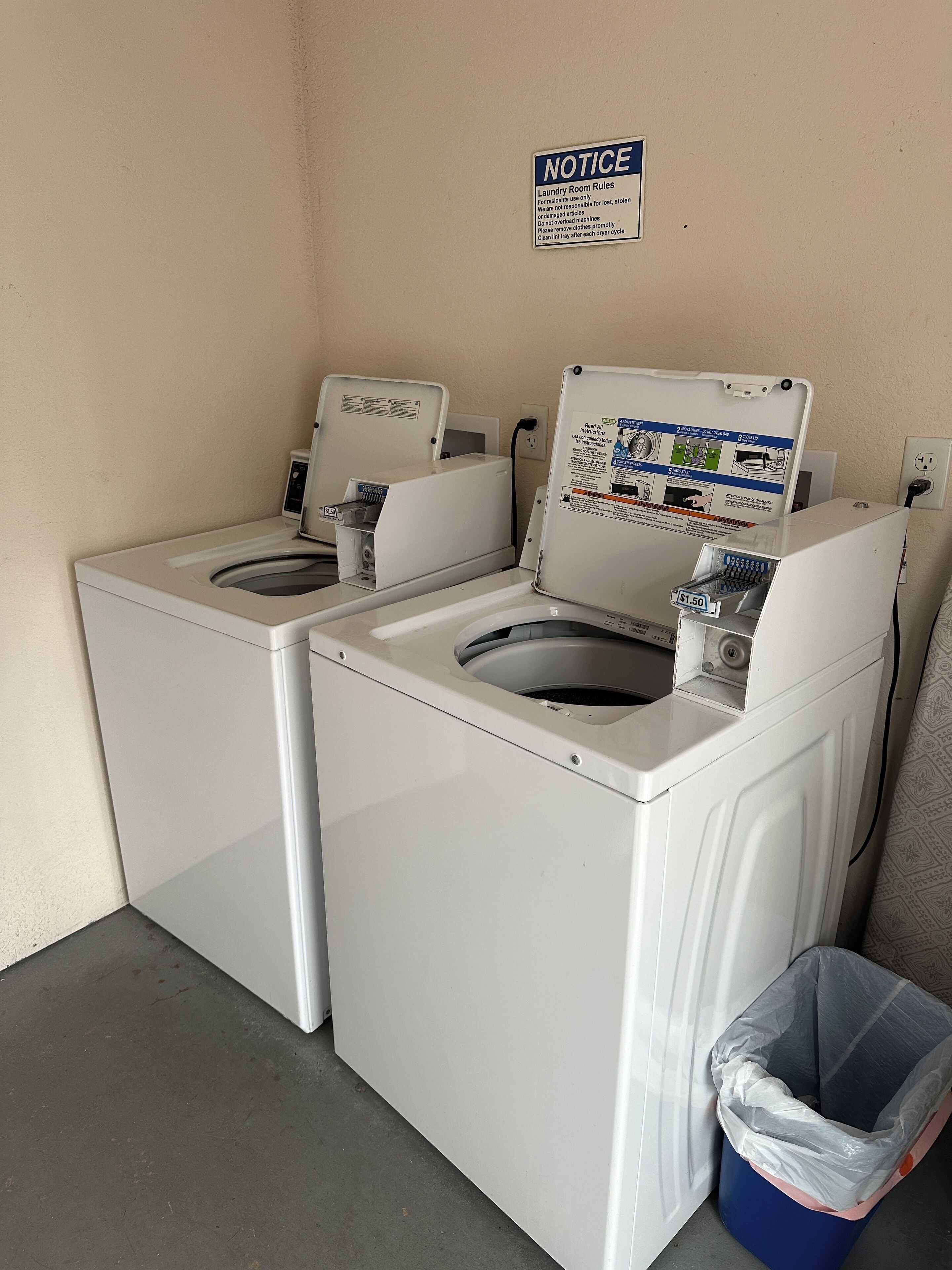 A laundry room with two washing machines, a notice sign, and a trash bin.