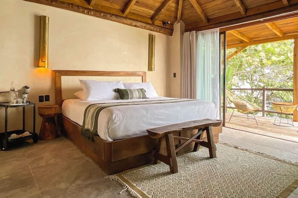A bedroom with a wooden bed, a bedside table, a bench, and a view of the outdoors.