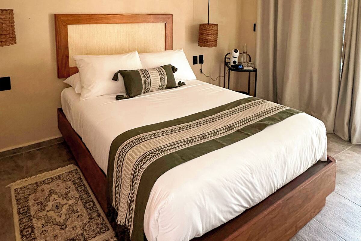 A large bed with a wooden frame, white bedding, and a patterned throw. A small table with a hairdryer and a bottle is visible in the corner.