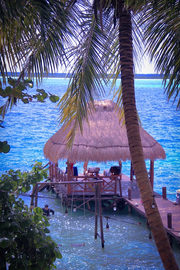 Relax on a wooden deck overlooking the calm water, surrounded by lush greenery and palm trees.