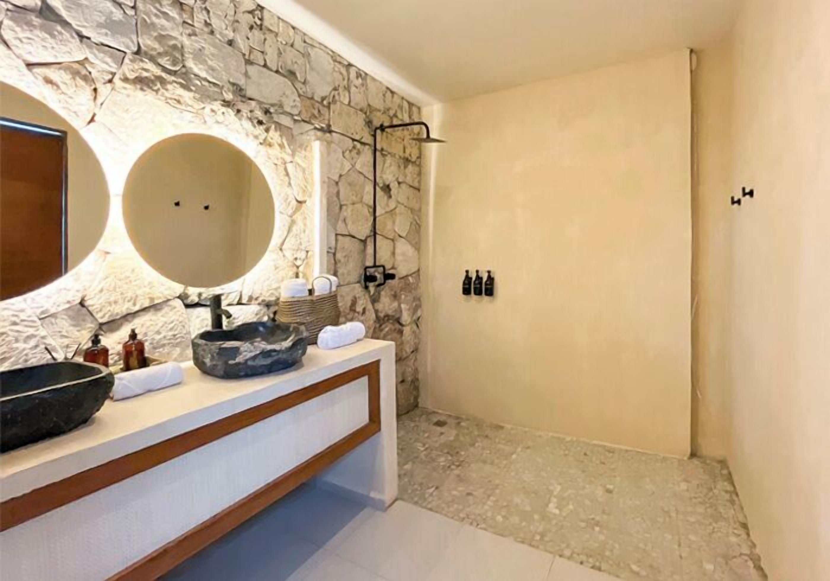 A bathroom with a stone wall, a round mirror, a large bowl sink, and a shower area.