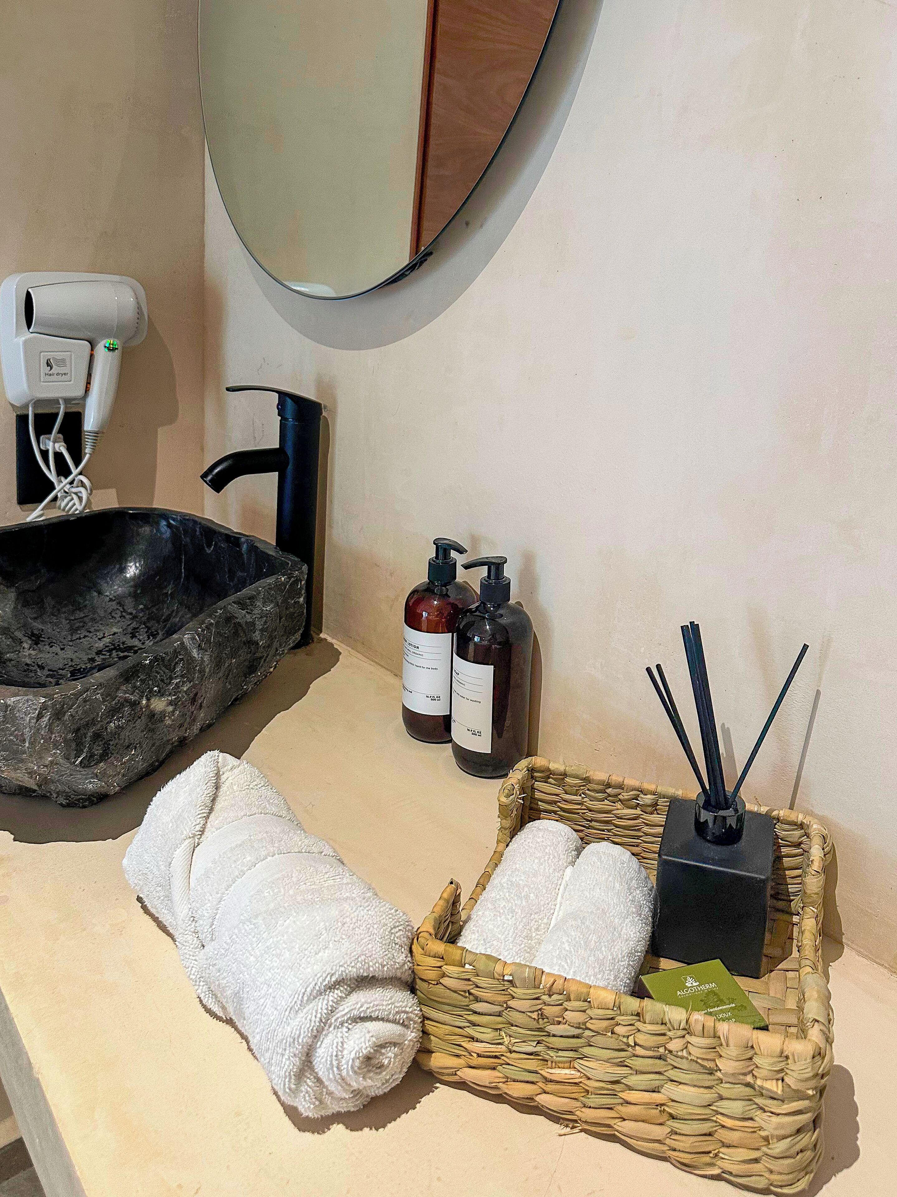 A bathroom with a black basin, two bottles of liquid soap, a black diffuser with reed sticks, and a woven basket containing two white towels.