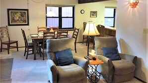 A living room with a sofa set, dining table, and chairs. There is a lamp, a clock, and a wall-mounted light fixture.