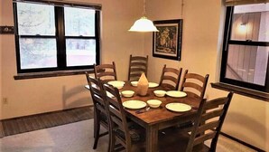 A dining area with a wooden table set for six, surrounded by six wooden chairs. There is a hanging light fixture above the table.