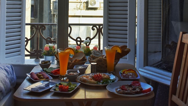 Ein Esstisch mit Frühstücksangeboten und Getränken, ein Fenster mit Rollläden und eine Ansicht von Gebäuden im Hintergrund.