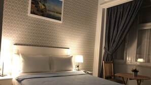Ein ordentlich bezogenes Bett mit blauer Tagesdecke, eine Nachttischlampe und ein Nachttisch mit Lampe. Ein kleiner Holztisch mit Topfpflanze steht neben dem Fenster.